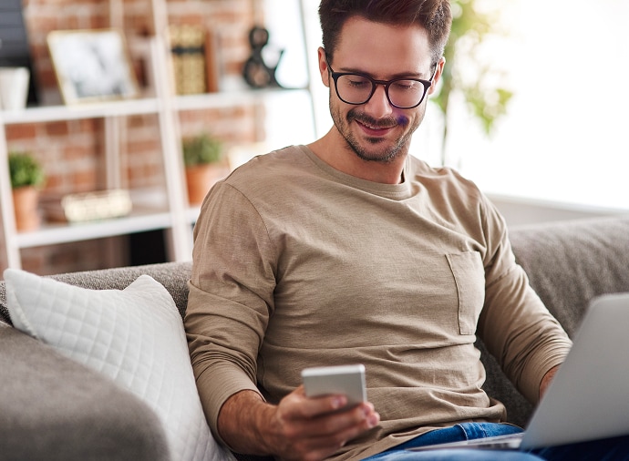 Homme assis sur un canapé regardant son téléphone, avec un ordinateur portable sur ses genoux